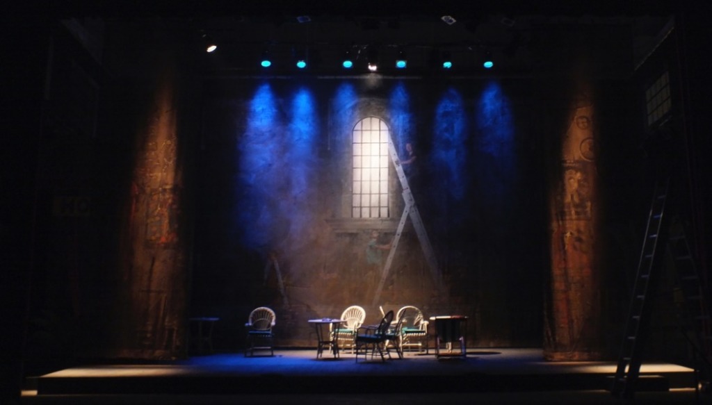 Wir sehen eine Theaterbühne im dunklen Scheinwerferlicht, kurz vor der Vorstellung arbeitet noch ein Bühnenarbeiter auf der Leiter.