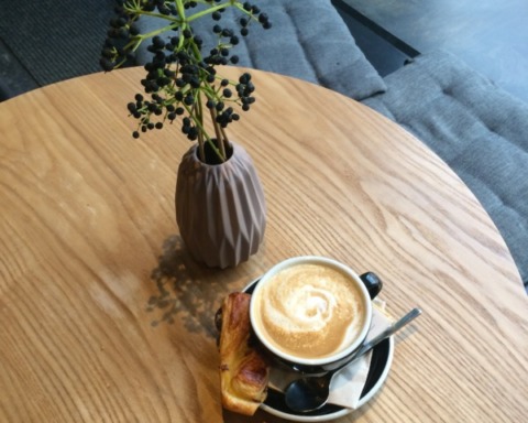 Auf einem Tisch steht eine Tasse Cappuccino. Dazu sieht man noch eine dekorative, moderne Vase mit einem beerenartigen Pflanzestiel. Der Tisch ist in einem Café.
