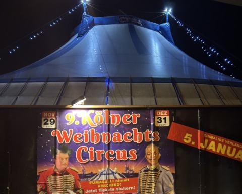 Ansicht des Kölner Weihnachtszirkus 2024 mit einem Plakat im Vordergrund und dem Zirkuszelt im Hintergrund. Alles kurz vor der Abend-Vorstellung.