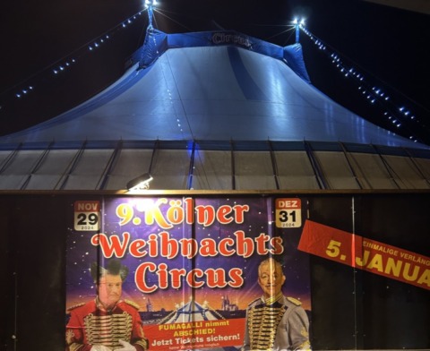 Ansicht des Kölner Weihnachtszirkus 2024 mit einem Plakat im Vordergrund und dem Zirkuszelt im Hintergrund. Alles kurz vor der Abend-Vorstellung.