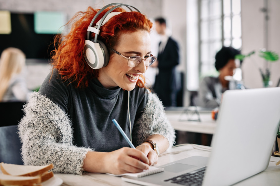 Fröhliche Unternehmerin, die an einem Computer arbeitet und sich Notizen in ihrem Notizblock macht, während sie im Büro Musik über Kopfhörer hört Es gibt Menschen im Hintergrund.