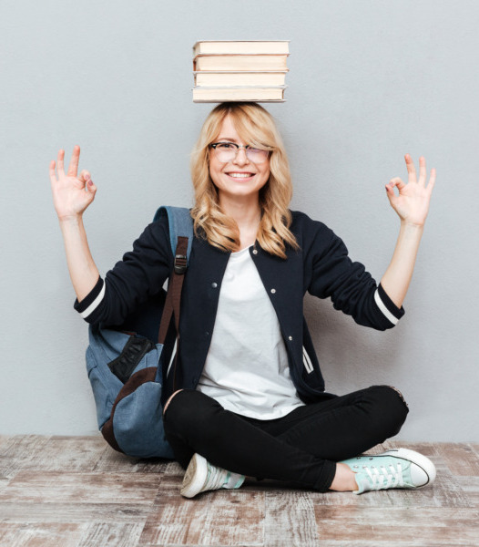 Picture of happy young woman student wearing glasses with backpack sitting over grey wall. Looking at camera holding books on head.