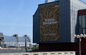 Wir sehen ein Außenplat der Ausstellung Yayoi Kusama an eine Hausmauer.
