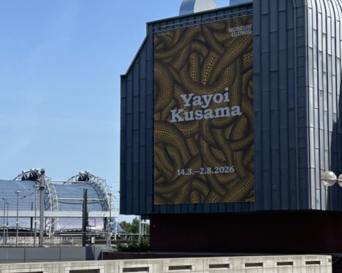 Wir sehen ein Außenplat der Ausstellung Yayoi Kusama an eine Hausmauer.