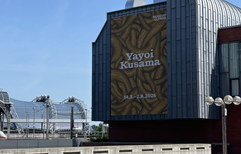 Wir sehen ein Außenplat der Ausstellung Yayoi Kusama an eine Hausmauer.