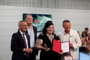 Wie sehen Lena Schätte auf der Bühnen mit drei Herren. Sie hält ihre Preisauszeichnungen in den Händen vor sich