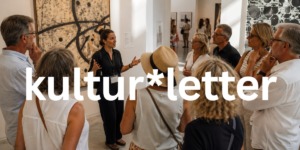 Eine Gruppe von acht Erwachsenen nimmt an einer Führung durch eine moderne Kunstausstellung teil. Eine Museumsführerin erklärt vor einem großformatigen abstrakten Gemälde ein Kunstwerk, während die Besucher aufmerksam zuhören. Der helle Ausstellungsraum ist lichtdurchflutet und vermittelt eine ruhige, sommerliche Atmosphäre.