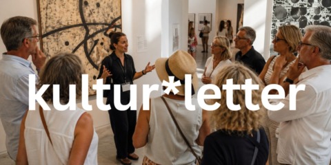 Eine Gruppe von acht Erwachsenen nimmt an einer Führung durch eine moderne Kunstausstellung teil. Eine Museumsführerin erklärt vor einem großformatigen abstrakten Gemälde ein Kunstwerk, während die Besucher aufmerksam zuhören. Der helle Ausstellungsraum ist lichtdurchflutet und vermittelt eine ruhige, sommerliche Atmosphäre.